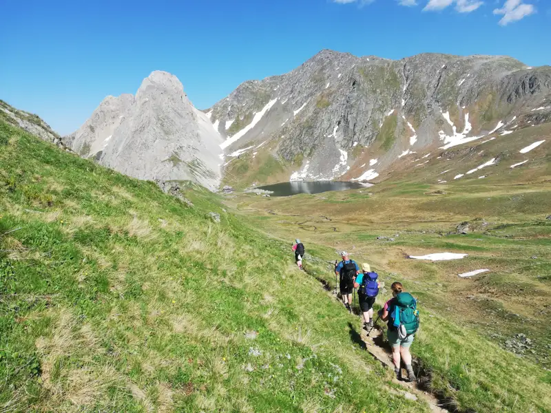 Wanderer auf einem Bergpfad inmitten einer Berglandschaft mit See und Gipfeln.