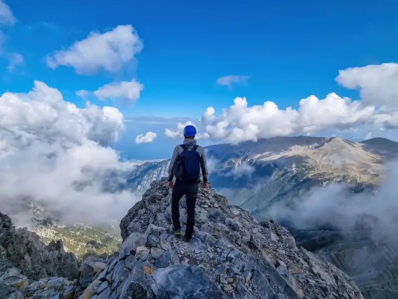 Wanderer auf einem Berggipfel mit Blick auf eine Berglandschaft und Wolken.