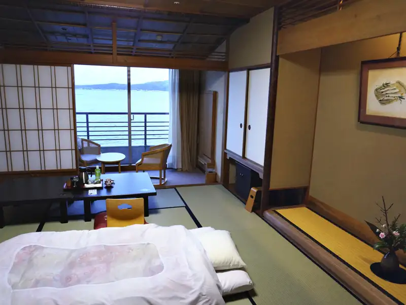 Innenansicht eines traditionellen japanischen Hotelzimmers mit Meerblick durch die Balkontür. Tatami-Matten, niedriger Tisch und traditionelle Schiebetüren sind sichtbar.