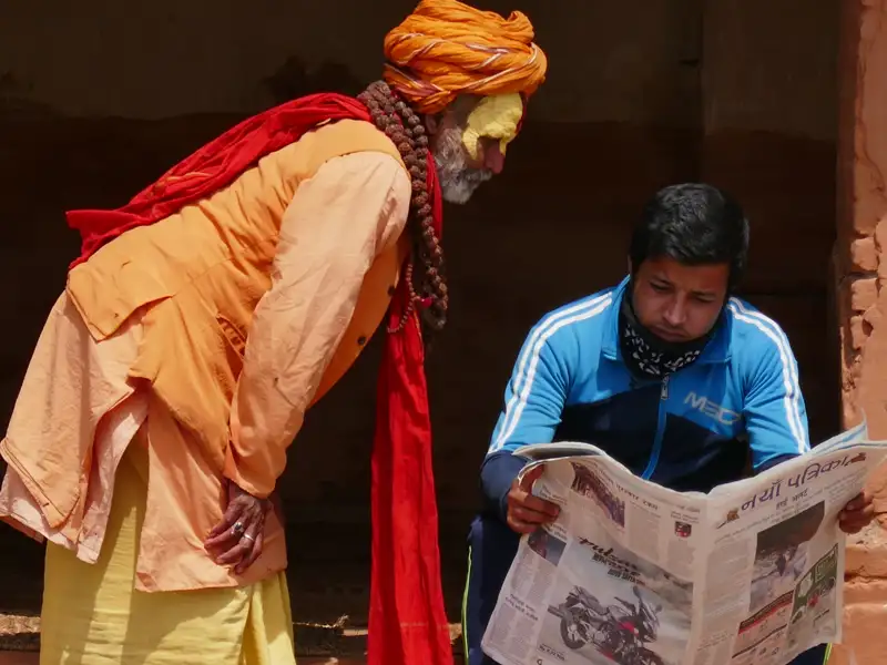 Ein Mann liest eine Zeitung, während ein Sadhu in traditioneller Kleidung ihm über die Schulter blickt. Die Szene spielt vermutlich in Nepal oder Indien und zeigt eine Begegnung zwischen traditionellem und modernem Leben.