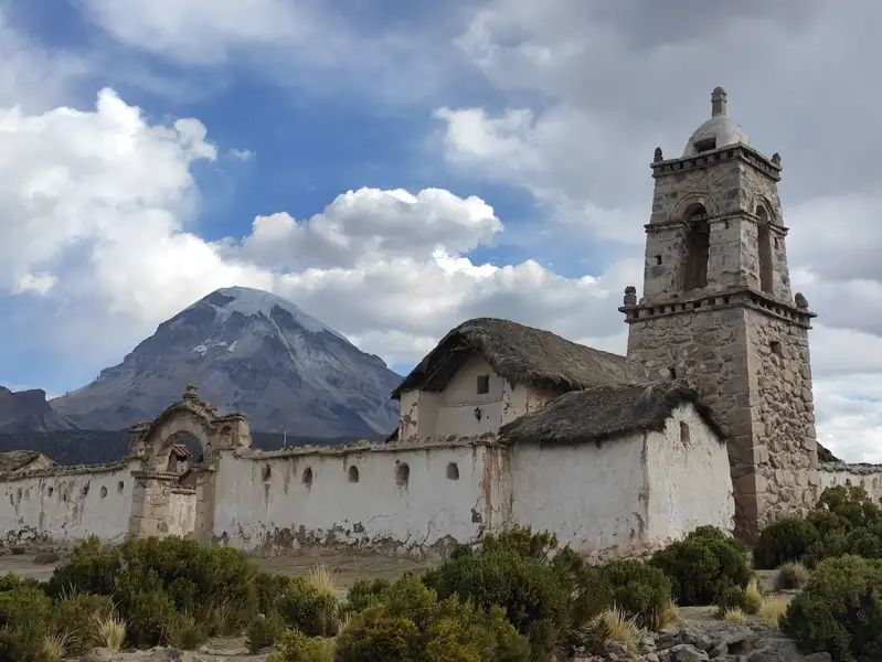 Alte Kirche mit Glockenturm und Strohdach vor majestätischem, schneebedecktem Berg.