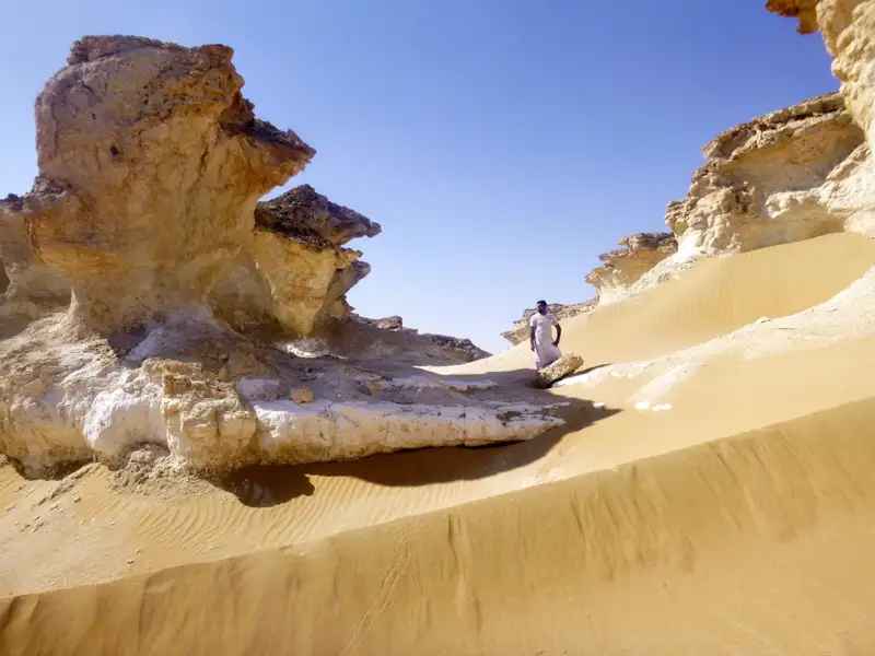 Person in traditioneller Kleidung inmitten von Wüstenfelsformationen und Sanddünen.