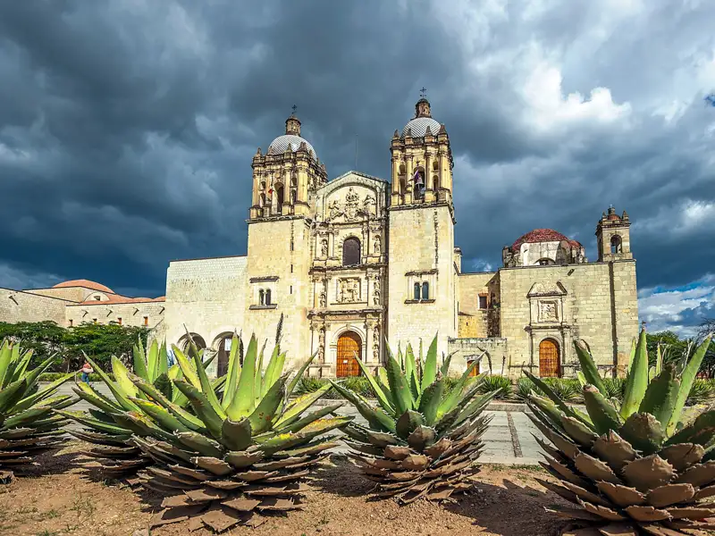 Historische Kirche mit zwei Türmen und Agaven im Vordergrund unter bewölktem Himmel.