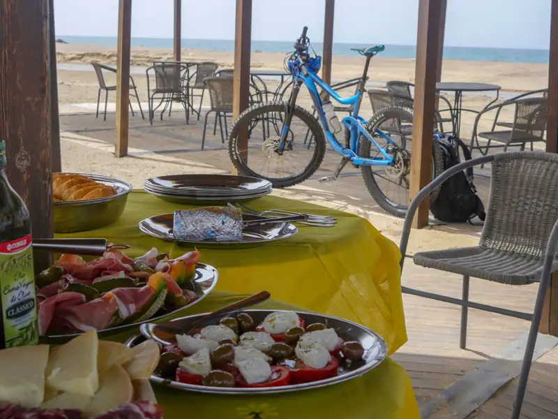 Mittagessen am Strand während einer Radtour. Ein blaues Mountainbike steht neben einem Tisch mit Essen, im Hintergrund ist das Meer zu sehen.