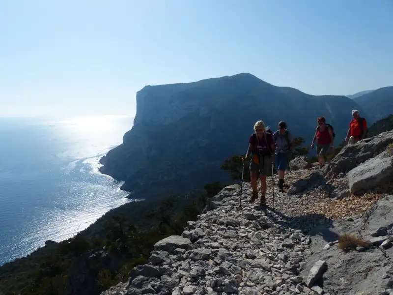 Wanderer mit Wanderstöcken auf einem steinigen Küstenpfad. Das Meer und eine steile Klippe sind im Hintergrund sichtbar.
