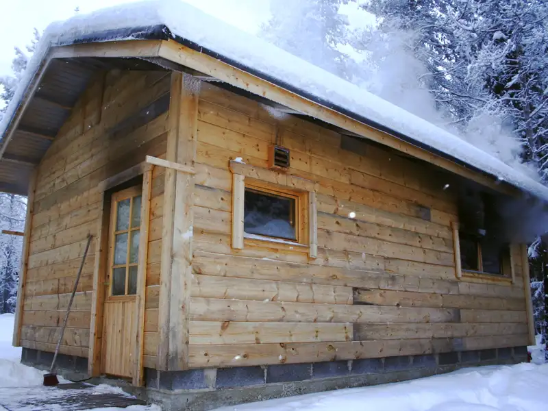 Eine verschneite Holzhütte mit aufsteigendem Rauch, der auf eine Wärmequelle im Inneren hindeutet.