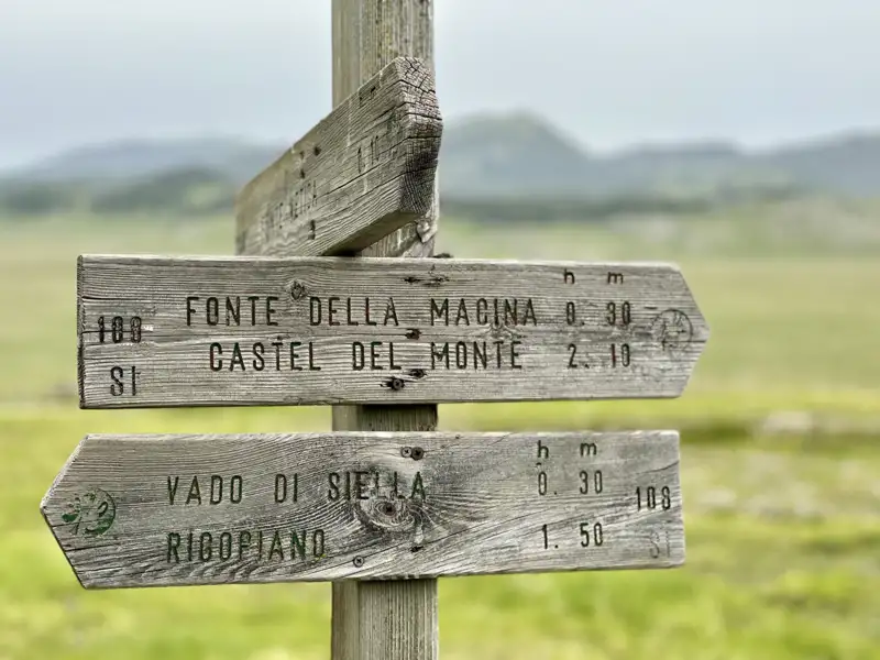 Wegweiser mit Wanderzeiten zu verschiedenen Zielen, darunter Fonte della Macina, Castel del Monte, Vado di Siella und Rigopiano.