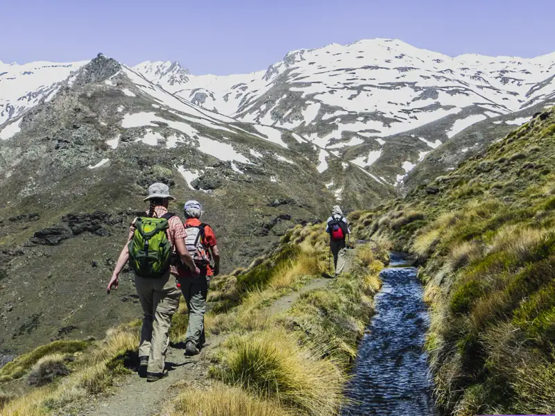 Wanderer auf einem Bergpfad mit schneebedeckten Gipfeln im Hintergrund. Ein Bach begleitet den Weg.