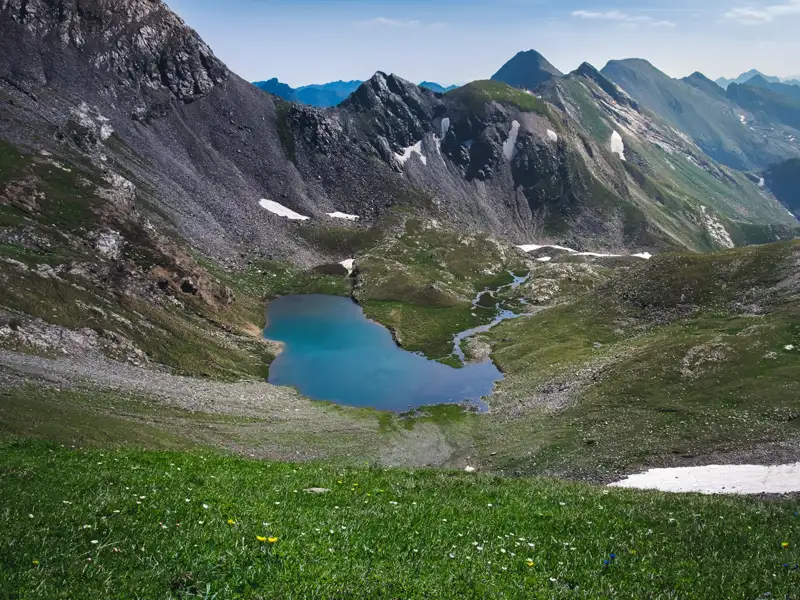 Blick auf einen blauen Bergsee inmitten der Alpen. Steile, felsige Hänge und Schneefelder umgeben den See. Eine grüne Wiese mit Wildblumen liegt im Vordergrund.
