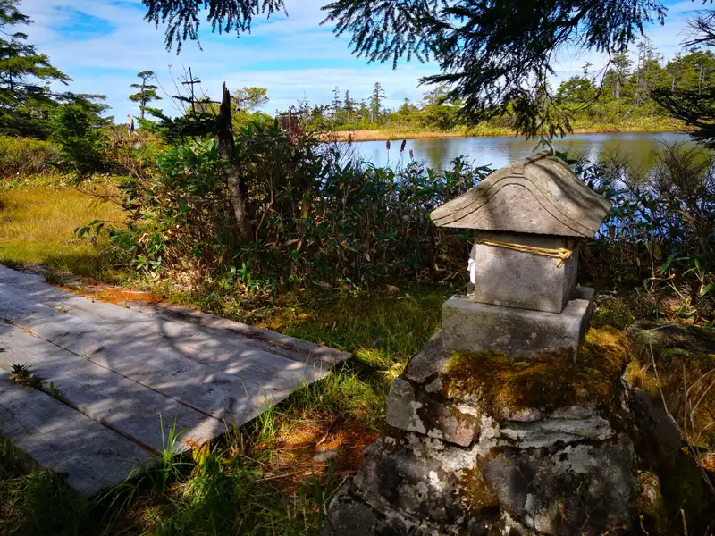 Steinlaterne am Ufer eines Bergsees, Holzsteg und Wanderer im Hintergrund.
