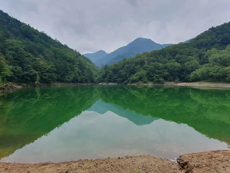 Blick auf einen smaragdgrünen See, umgeben von bewaldeten Hügeln unter einem bewölkten Himmel.