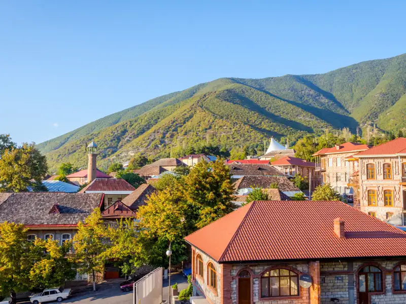 Panoramablick auf eine Stadt mit traditioneller Architektur, einer Moschee und einem bewaldeten Berghang im Hintergrund.