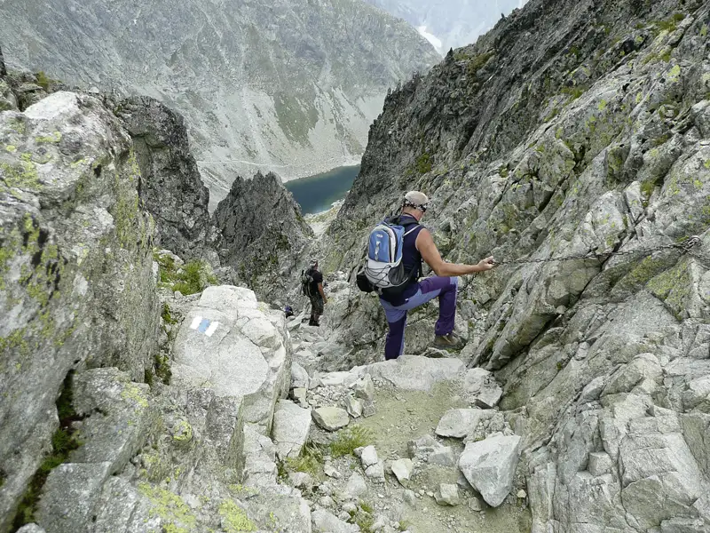 Wanderer auf einem anspruchsvollen Bergpfad mit Ketten und Blick auf einen Bergsee.