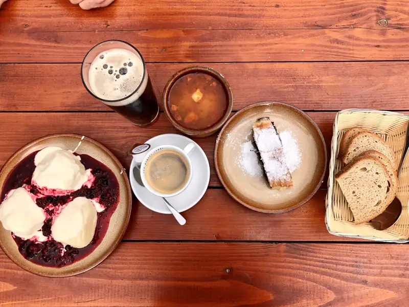 Eine Auswahl an traditionellen Speisen und Getränken, darunter Dampfnudeln, Mohnstrudel, Suppe und Brot, zusammen mit einem dunklen Bier und Kaffee.