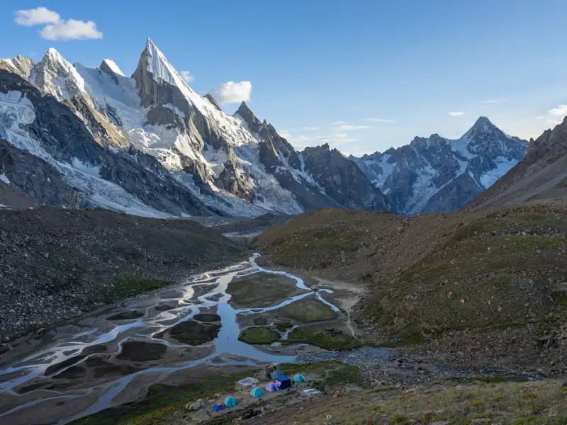 Basislager der Reise in einer hochalpinen Umgebung. Zelte stehen am Ufer eines sich verzweigenden Flusses, umgeben von schneebedeckten Gipfeln.