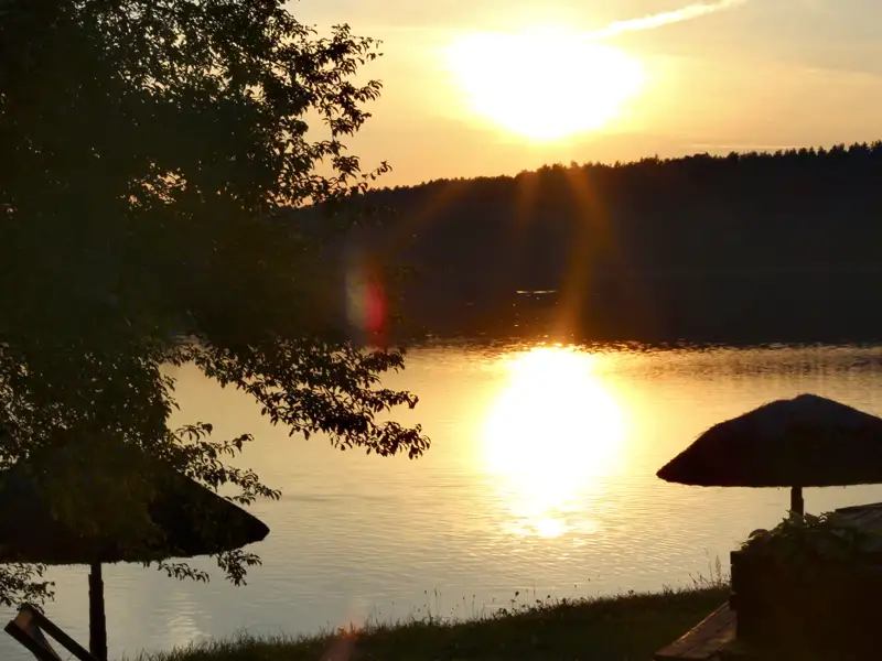 Sonnenuntergang über einem See, gesehen von einem Ufer mit Sonnenschirmen.