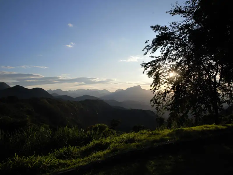 Sonnenuntergang hinter den Bergen, gesehen von einem Aussichtspunkt mit Gras und einem Baum im Vordergrund.