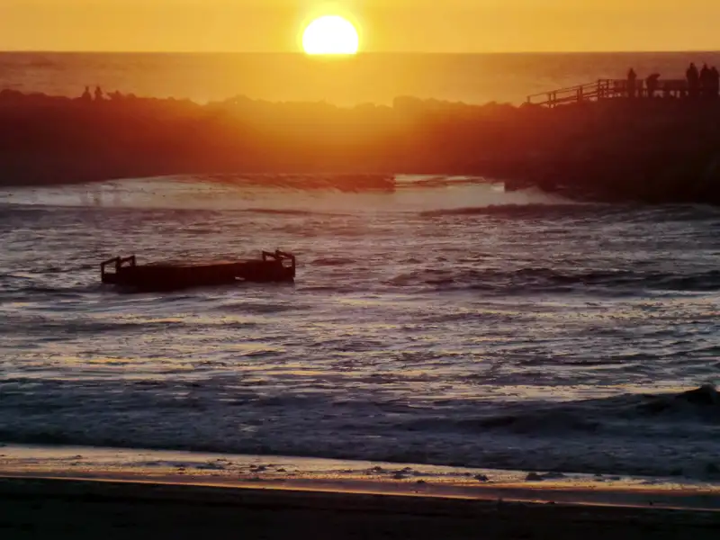 Sonnenuntergang über dem Meer mit einem teilweise zerstörten Steg im Vordergrund und Menschen, die auf einer Plattform den Sonnenuntergang beobachten.