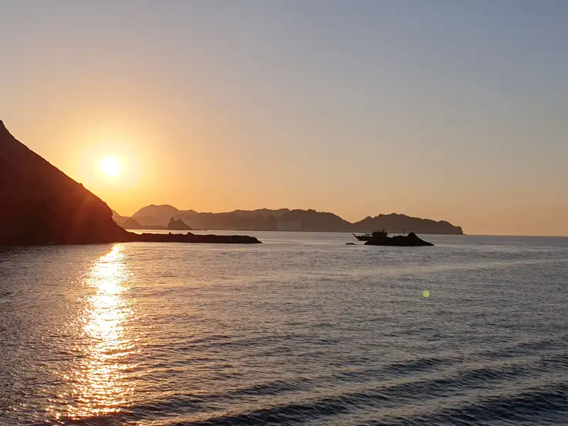 Sonnenuntergang über dem Meer mit Silhouette eines Bootes und Bergen.