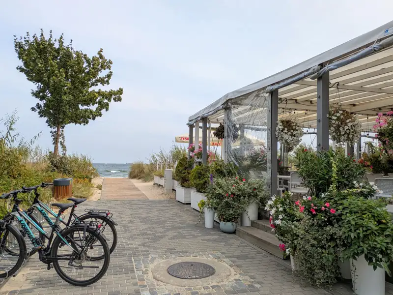 Strandrestaurant mit überdachter Terrasse und blühenden Blumen. Zwei Fahrräder stehen am Eingang. Ein Weg führt zum Strand.