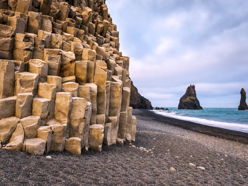 Geometrische Basaltsäulenformationen am schwarzen Sandstrand mit Blick auf die charakteristischen Felsnadeln im Meer.