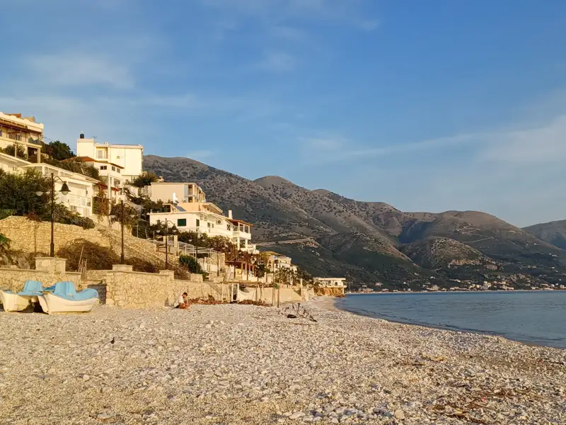 Blick auf einen Kiesstrand mit Booten, Häusern am Hang und Bergen im Hintergrund.