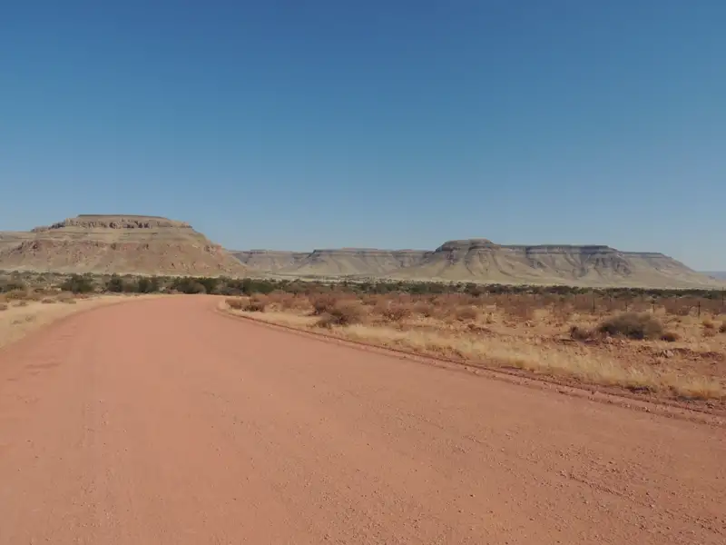 Rote Schotterstraße in einer trockenen Landschaft mit Tafelbergen im Hintergrund.