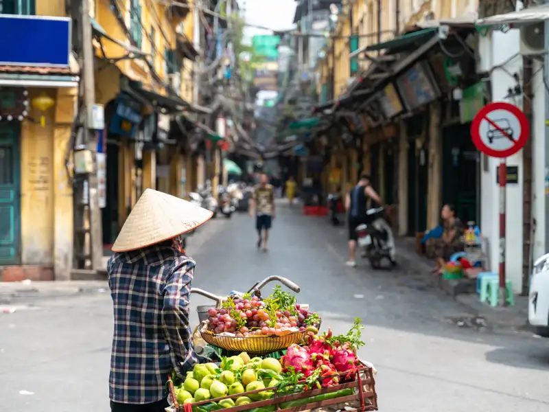 Straßenverkäuferin mit Strohhut und Fahrrad voller frischer Früchte in Hanoi.