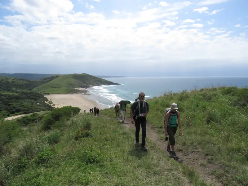 Wanderer mit Rucksäcken auf einem Küstenpfad mit Blick auf den Strand und das Meer.
