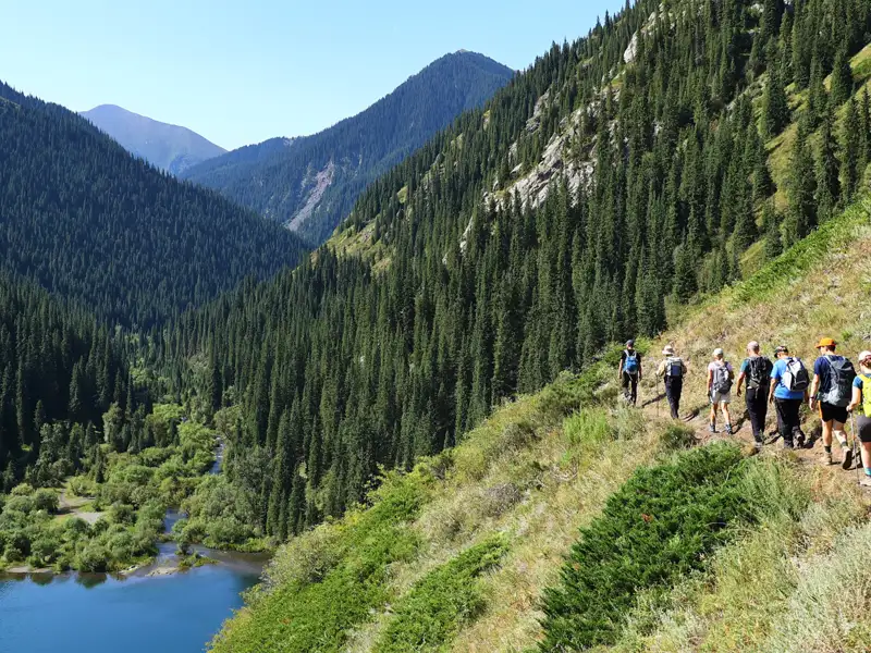 Wandergruppe auf einem Bergpfad inmitten einer bewaldeten Berglandschaft mit Blick auf einen Fluss.