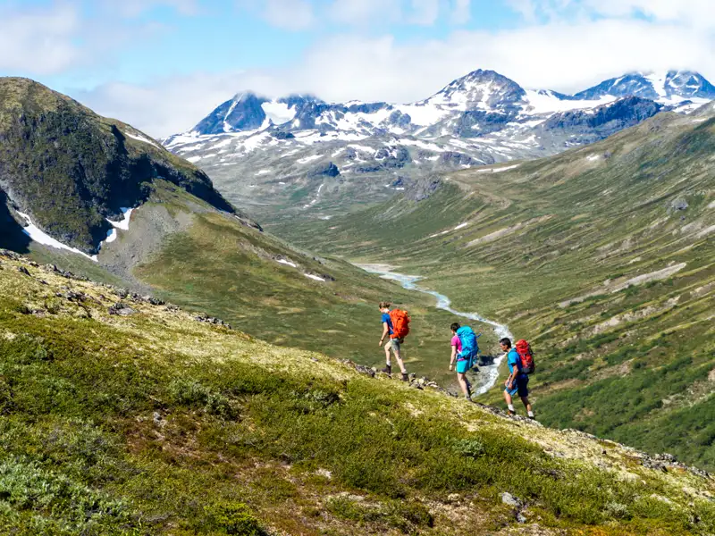 Drei Wanderer mit Rucksäcken besteigen einen grünen Berghang. Im Hintergrund erstreckt sich ein Tal mit einem Fluss und schneebedeckten Bergen.