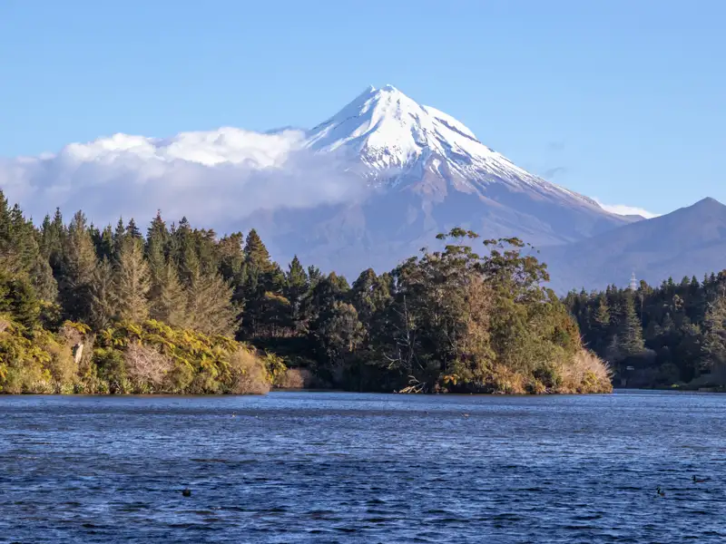 Panoramablick auf einen schneebedeckten Berg vom Seeufer aus, umgeben von Bäumen.