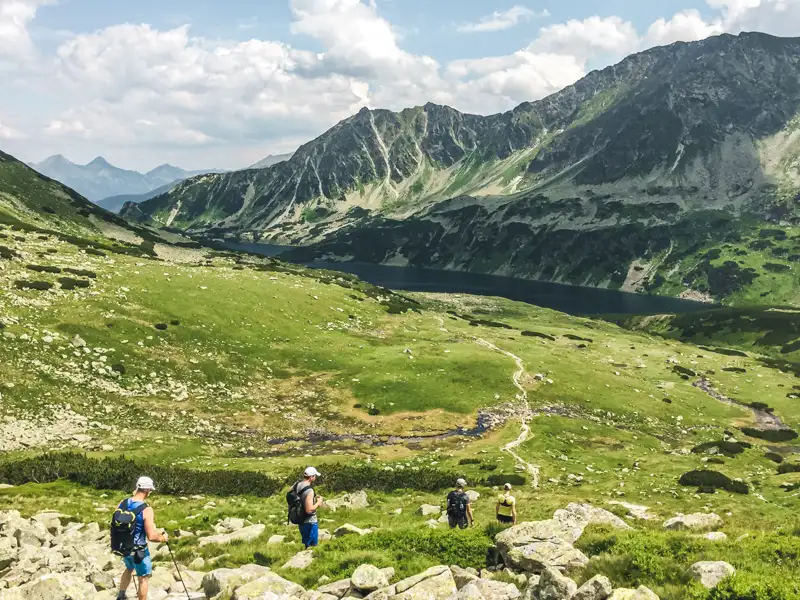 Wanderer mit Rucksäcken und Wanderstöcken auf einem Bergpfad in Richtung eines Sees und hoher Berge.
