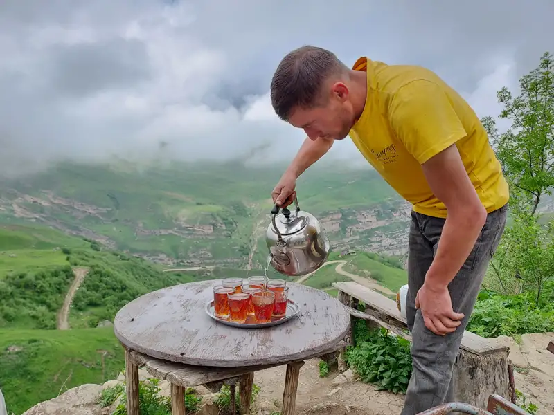 Ein Reisender schenkt Tee in Gläser ein, mit einem malerischen Blick auf das Tal im Hintergrund.