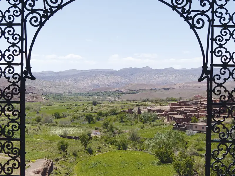 Blick durch ein verziertes Tor auf ein grünes Tal mit einem Dorf und Bergen im Hintergrund.