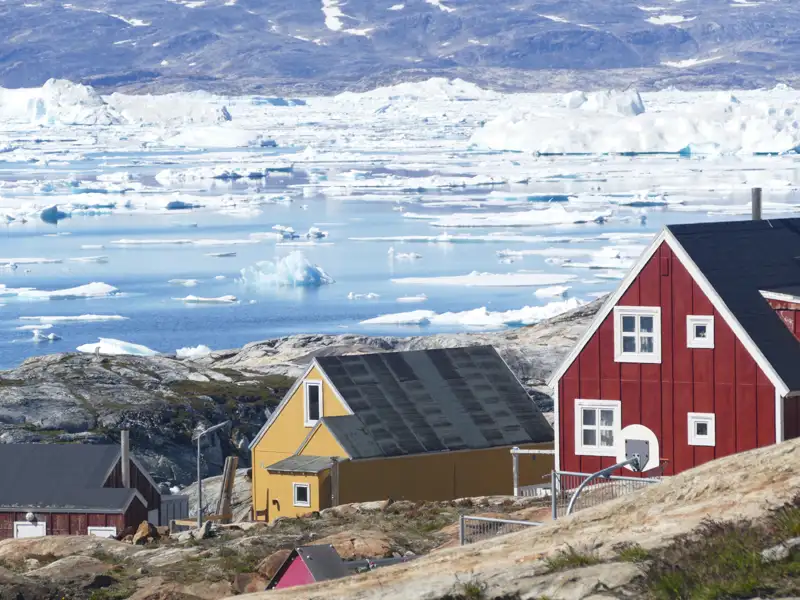 Traditionelle Häuser in einer grönländischen Küstensiedlung mit Blick auf das eisbedeckte Meer und zahlreiche Eisberge.