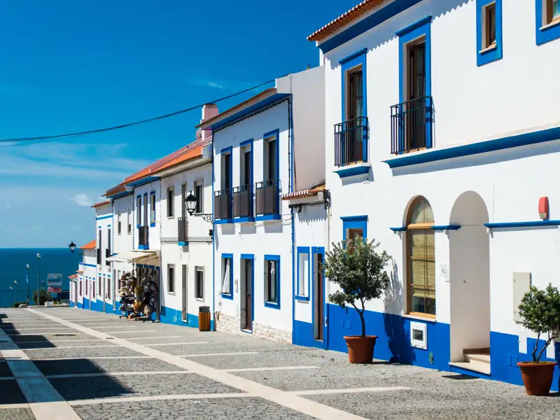 Weiß-blaue Häuser säumen eine Straße in einer Küstenstadt. Das Meer ist im Hintergrund sichtbar.