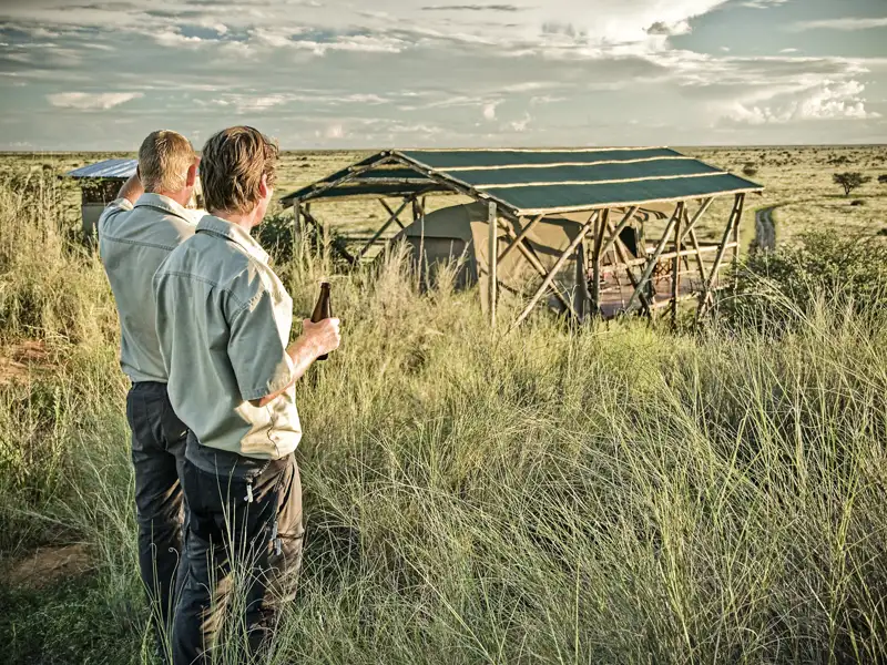 Zwei Reisende genießen den Blick auf ihre Unterkunft, eine Safari-Lodge inmitten der afrikanischen Savanne.