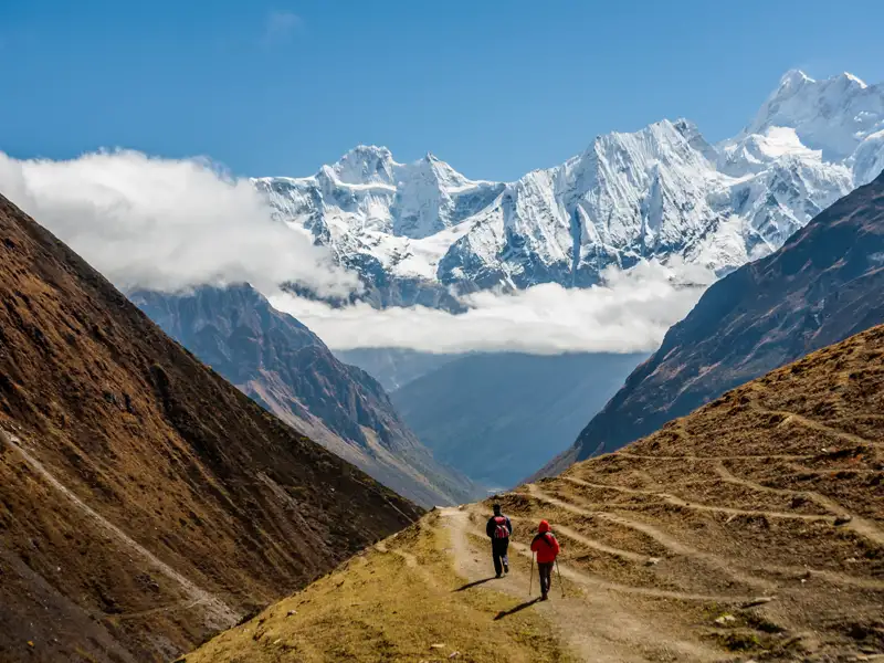 Zwei Wanderer auf einem Bergpfad im Himalaya. Im Hintergrund sind schneebedeckte Gipfel zu sehen.
