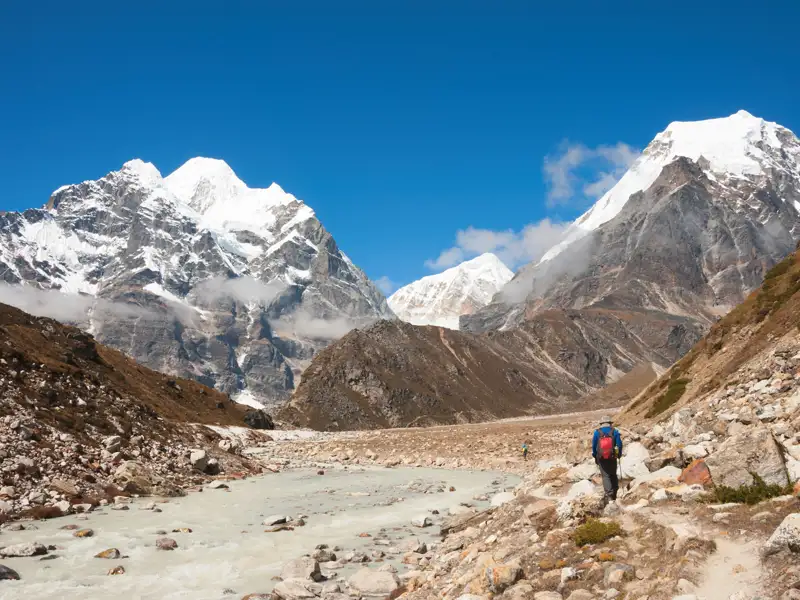 Wanderer auf einem Bergpfad neben einem Gletscherfluss, umgeben von schneebedeckten Gipfeln.