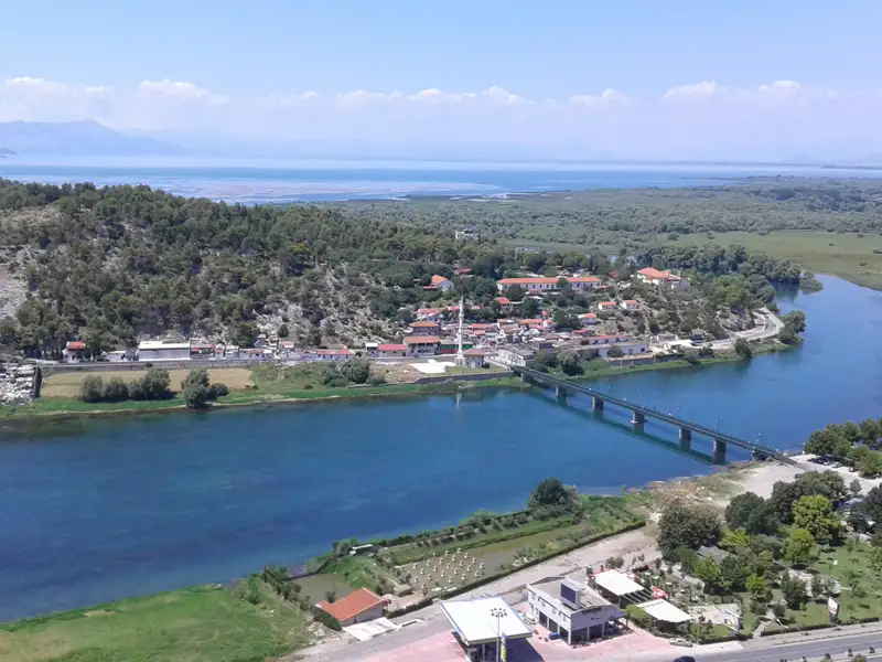 Luftbild eines Dorfes an einem Fluss mit Brücke und See im Hintergrund.