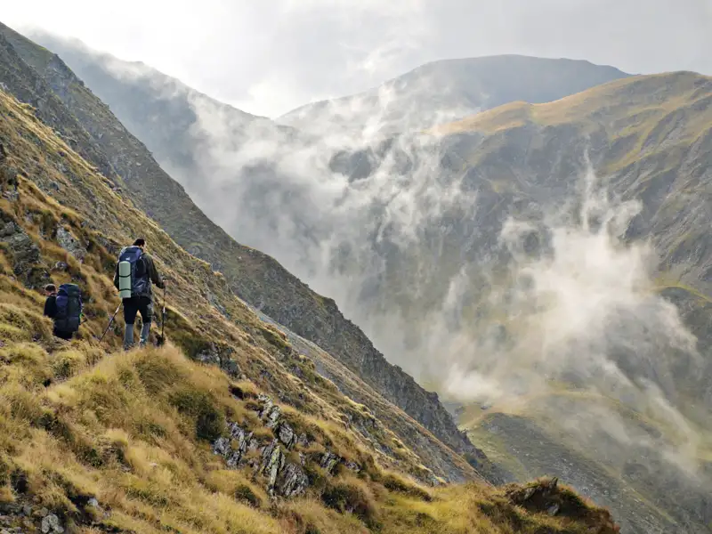 Zwei Wanderer mit Trekkingstöcken und Rucksäcken auf einem Bergpfad. Nebel steigt im Tal auf.
