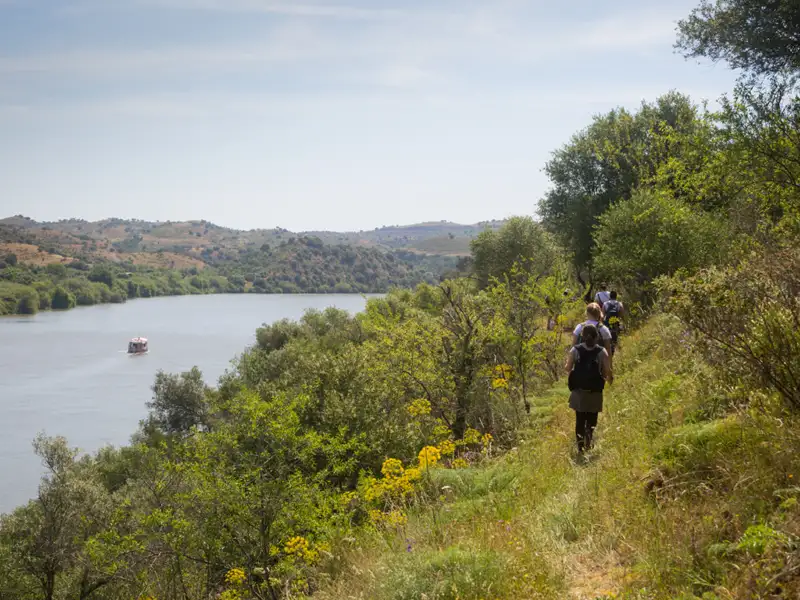 Wanderer auf einem Pfad entlang des Flusses. Ein Boot fährt im Hintergrund auf dem Wasser.