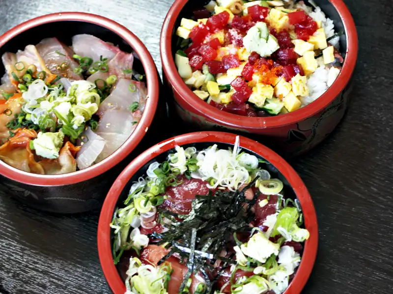 Drei Schalen mit verschiedenen Fischgerichten, darunter roher Fisch, möglicherweise Thunfisch oder ein ähnlicher Fisch, sowie ein Gericht mit Reis und verschiedenen Toppings.