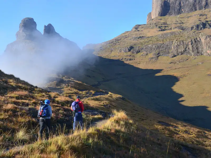 Zwei Wanderer auf einem Bergpfad.  Felsige Gipfel und Nebel sind im Hintergrund sichtbar.
