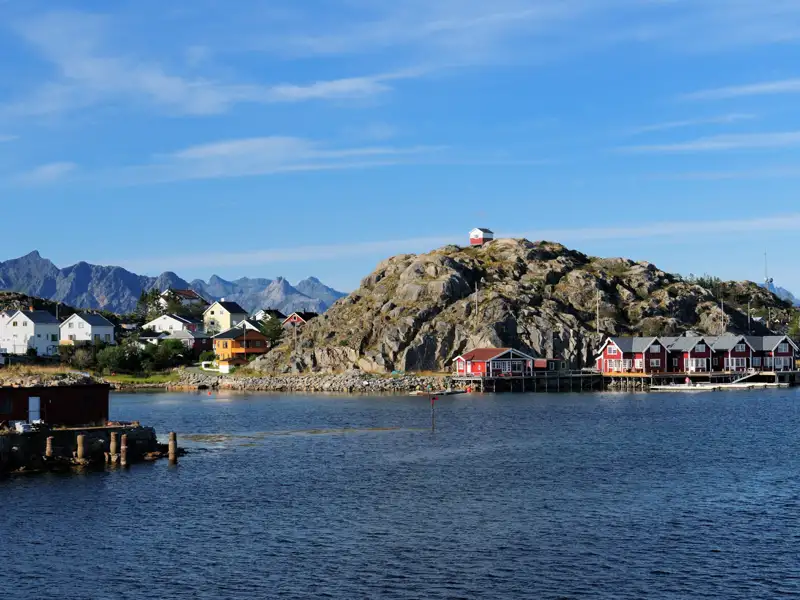 Buntes Dorf an der Küste mit felsiger Insel und Bergen im Hintergrund.