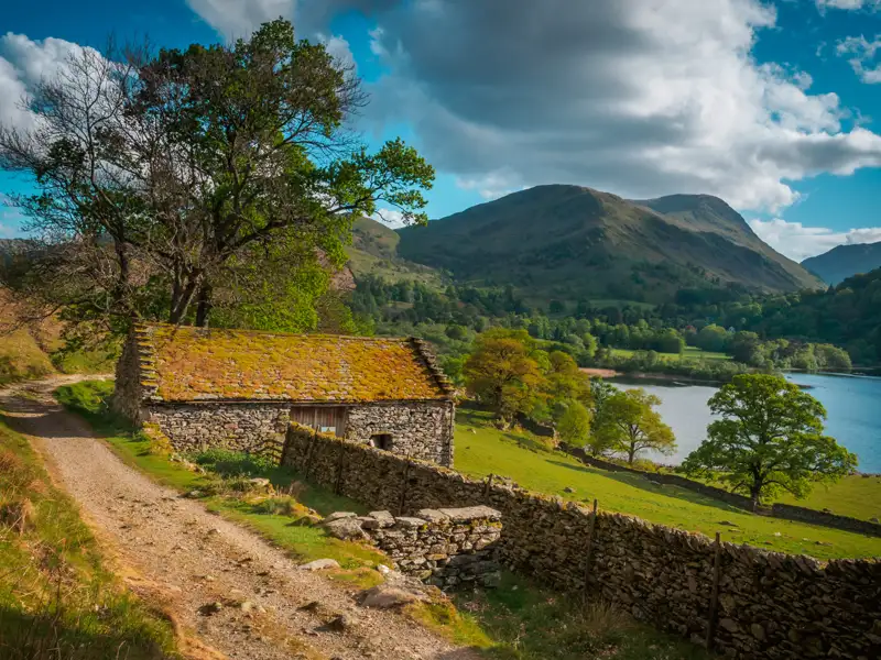 Steinernes Gebäude mit bemoostem Dach in einer Landschaft mit See und Bergen.