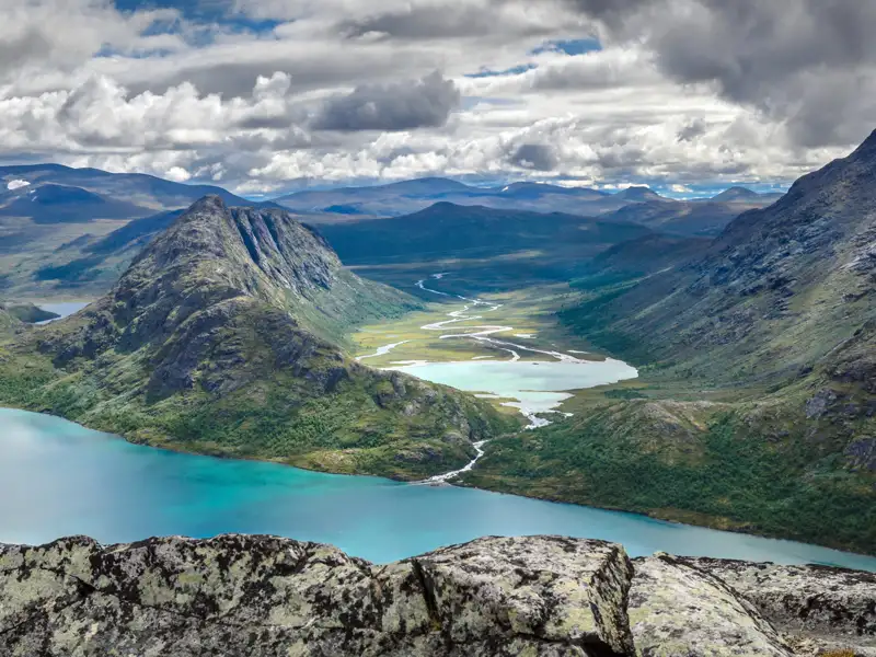 Aussicht von einem Felsvorsprung auf einen türkisfarbenen See inmitten einer bergigen Landschaft.