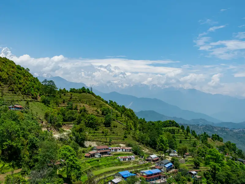 Panoramablick auf ein Bergdorf mit terrassierten Feldern und den schneebedeckten Himalaya-Gipfeln im Hintergrund.
