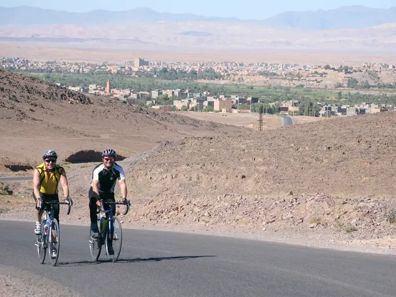 Zwei Radfahrer auf einer Radtour in einer Wüstenlandschaft. Im Hintergrund ist eine Stadt sichtbar.